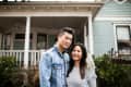 Couple smiling in front of a light blue house with a white porch and greenery.