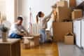 Couple unpacking boxes in a kitchen, with a man kneeling and a woman reaching for items on stacked boxes.