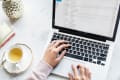 Hands typing on a laptop with an open email inbox, next to a cup of tea and a decorative pineapple.