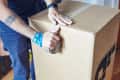 Person sealing a large cardboard box with packing tape, wearing a smartwatch and colorful wristbands.