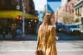 Woman in a yellow floral dress crossing a city street with blurred traffic and buildings in the background.