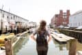 Person with backpack standing on a dock, overlooking a canal with boats and industrial buildings.