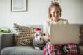 Woman in festive headband using laptop on sofa with dog in Santa hat and sweater.