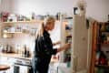 Woman in a kitchen opening a refrigerator, surrounded by shelves with jars, bottles, and kitchen items.