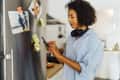 Woman in a kitchen using a smartphone, wearing headphones around her neck, with photos and sticky notes on the fridge.