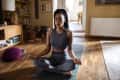 Woman meditating in a living room on a yoga mat, wearing a gray workout outfit, surrounded by books and decor.
