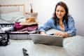 Young woman using laptop for booking airplane tickets for her vacation. Packed suitcase next to her