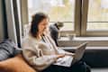 Young woman working at home remotely or online shopping using a laptop while sitting on the sofa. Her domestic cat is sitting next to her