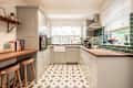 A general interior view of a victorian country style taupe coloured domestic kitchen, wooden worktop, green wall tiles, brass handles, neo-classical star shape patterned floor tiles, breakfast bar with wooden bar stools, butler sink, window with venetian blinds, black oven, ceramic induction hob