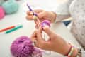 A close-up view of a woman’s hands as she skillfully crochets with purple yarn. The background includes colorful crochet hooks, balls of wool, and a pincushion, showcasing an organized and vibrant crafting environment. The bright workspace highlights her focus and creativity.