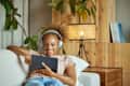 Smiling young woman watching movie on tablet PC. Female is wearing wireless headphones while using wireless technology. She is reclining on sofa at home.