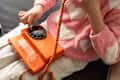 A teenage girl sits in a pink heart-patterned sweater, using a bright orange landline phone. The cozy bedroom setting conveys retro technology use and communication.