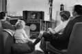 Family watching a small television in a living room, with a child sitting on cushions and a woman holding a magazine.
