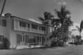 Two-story house with shutters and balcony, surrounded by palm trees and a grassy lawn.