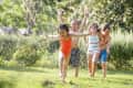 Children running through water sprinkler on a large lawn with trees, bushes in background
