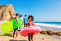 Photo of a young family of four enjoying a day at the beach; children are carrying inflatable beach toys and youngest daughter is bringing up the rear as she looks at the camera with a big smile.