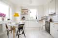 Bright kitchen with white cabinets, yellow pendant light, round table, mismatched chairs, and potted plant on windowsill.