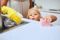 Child cleaning kitchen counter with pink sponge, adult wearing yellow gloves using spray bottle.