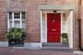 Red front door with brass knocker, flanked by white shutters, brick wall, and potted plants.