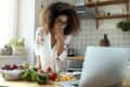 Woman in kitchen looking at laptop, surrounded by fresh vegetables and fruits on a wooden countertop.