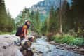 Hiker with backpack and trekking poles sitting by a forest stream, surrounded by tall trees and mountains.