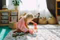 A young child in a pink dress plays on a soft rug, stacking colorful blocks beside toy baskets and a potted plant.