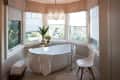 Freestanding bathtub in a bay window bathroom with a modern chair, plant, and chandelier.