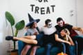 Four friends in Halloween costumes laughing on a blue sofa, with "Trick or Treat" banner and a potted plant nearby.