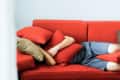 Person lying on a red sofa, partially covered with red and beige pillows, wearing blue shorts.