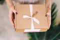 Hands holding a gift in a cardboard box with pink ribbon and tiny sprig of flowers