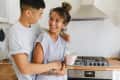 Couple smiling in a modern kitchen, one holding a pink mug near a stainless steel stove.