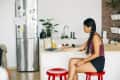 Woman with braided hair using a laptop at a kitchen counter, sitting on a red stool, with plants and a fridge in the background.