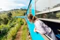 Woman leaning out of a blue train window, enjoying scenic green hills and wildflowers.