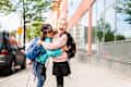 Two children with backpacks hugging and laughing on a city sidewalk near a glass building.
