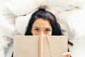 Woman lying on bed, holding a beige book, with only her eyes visible above it.