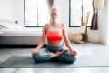 Woman meditating in a bright bedroom, sitting cross-legged on a yoga mat, wearing a red sports bra and gray pants.