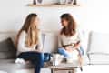 Two women sitting on a sofa, talking and smiling, with a wooden coffee table and shelf with photos in the background.