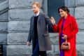 Man in gray coat and woman in red coat with brown handbag walking outside a stone building.