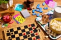 Board game setup with checkers, playing cards, dominoes, popcorn, gummy candies, and drinks on a wooden table.