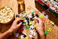 Hands assembling colorful puzzle pieces on a wooden table with a bowl of popcorn and a glass of iced drink nearby.