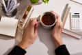 Hands holding a speckled mug of tea on a desk with a gold stapler, notepad, pens, and a calculator.
