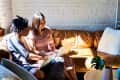 Two people sitting on a brown sofa, using a laptop, with sunlight streaming through a window.