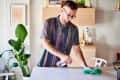 Man cleaning a white table with a spray bottle and cloth in a bright room with plants and shelves.