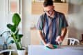 Man cleaning a table with a spray bottle and cloth, next to a large potted plant in a bright room.