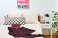 Bedroom with a wooden bed, checkered pillow, folded clothes, and a maroon sweater. White nightstand with lamp and plant.
