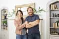 A smiling couple stands in a living room with bookshelves, plants, and a rustic mirror.