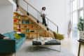 Modern living room with teal sofa, green chair, wooden bookshelf, and a person descending a staircase.