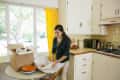 Woman unpacking dishes in a kitchen with yellow curtains, white cabinets, and a countertop with a toaster oven and kettle.