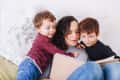 Woman reading a book to two young boys on a bed with floral pillows.
