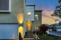 Modern two-story house exterior with garage, lit entrance, and landscaped garden at sunset.